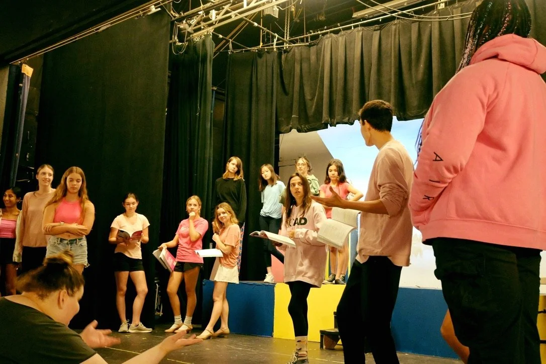 A group of young students on stage during a rehearsal or class, with some standing on risers and others in front, holding scripts or notebooks, and one girl in front appears to be reading aloud.