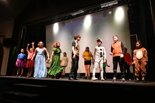 A group of children performing on a stage during a theatrical play, with some in costume and others standing in the background.