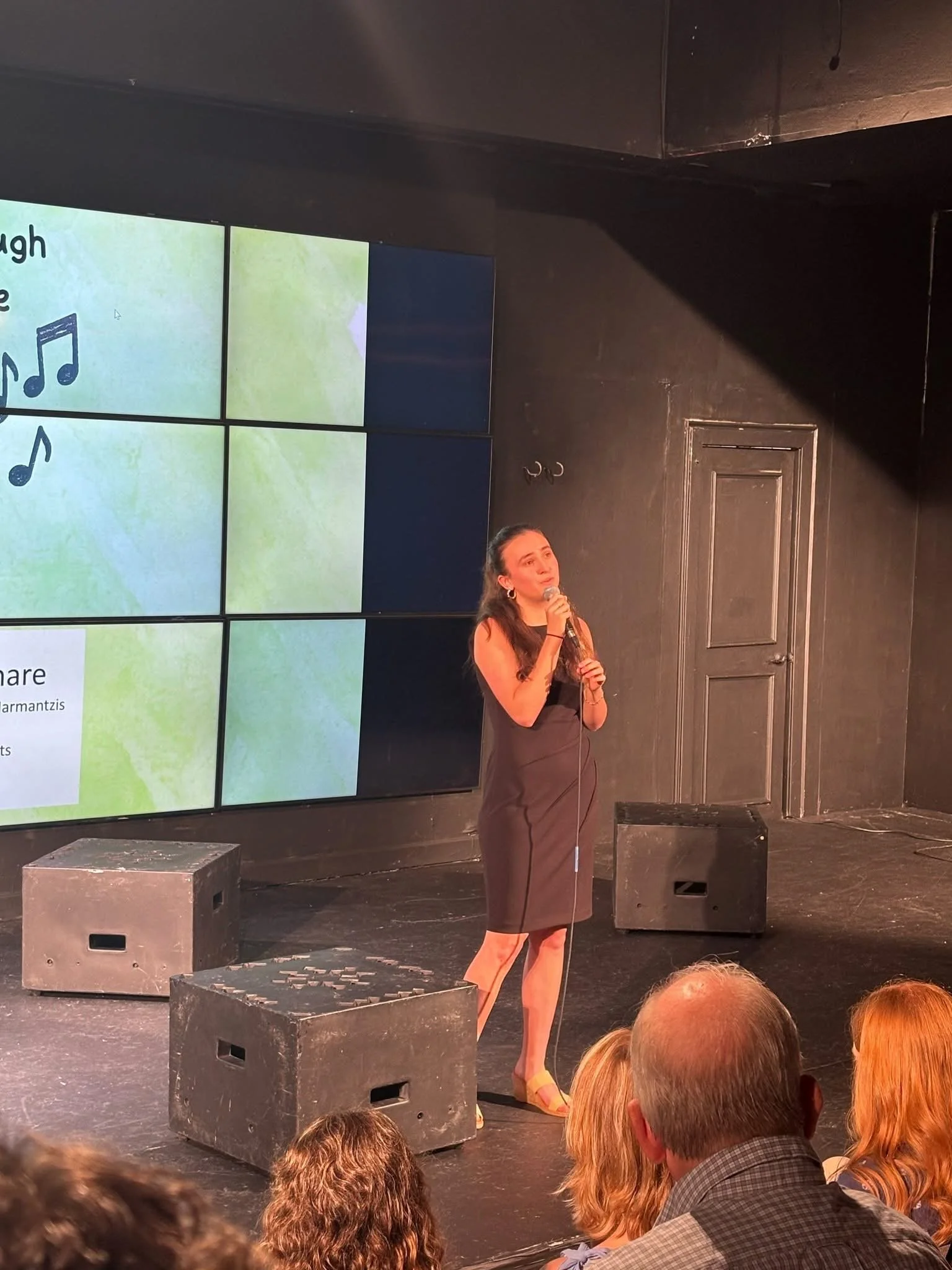 A woman stands on a stage holding a microphone and speaking to an audience, with a large screen behind her displaying lyrics and musical notes.