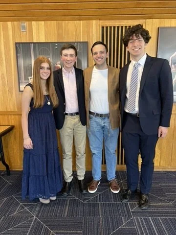 Four young adults standing together indoors, dressed in semi-formal attire, smiling for the camera.