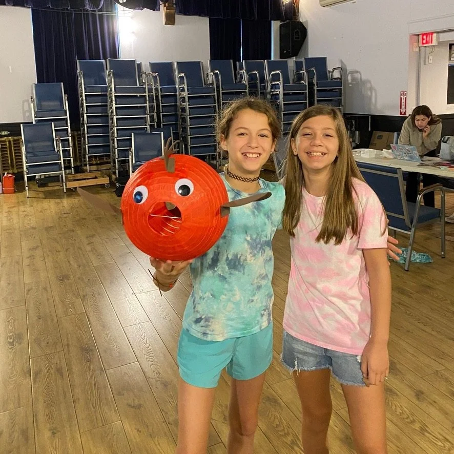 Two girls smiling inside a room with stacked chairs in the background. One girl is holding a pumpkin-shaped lantern with a surprised face, and the other girl is standing beside her with her arm around. The room has wood flooring and a woman sitting a