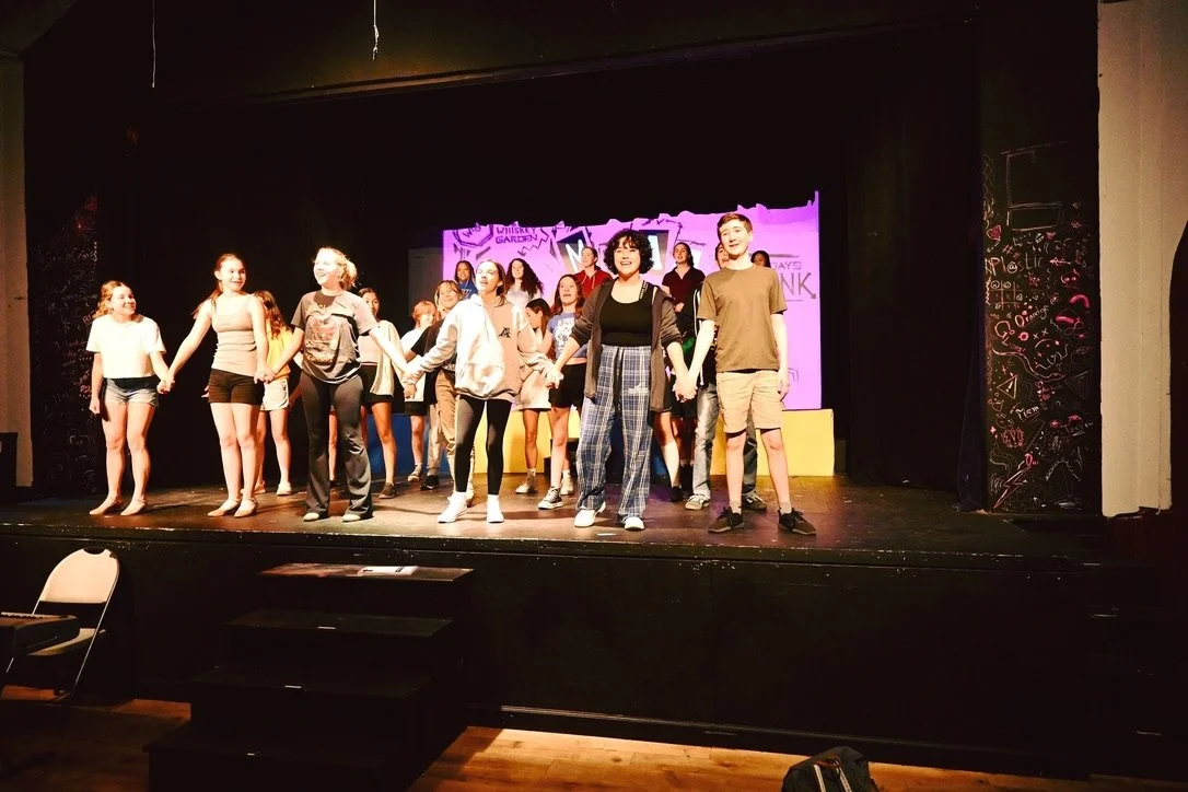 Group of children and teenagers performing a play on stage, holding hands, with a colorful backdrop.