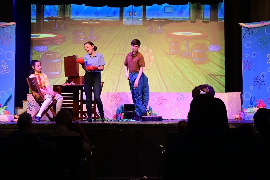 Children performing a play on stage with "SpongeBob SquarePants" theme, featuring a girl sitting on a chair, girl holding a pot, boy with a mop, and background of an animated underwater scene.
