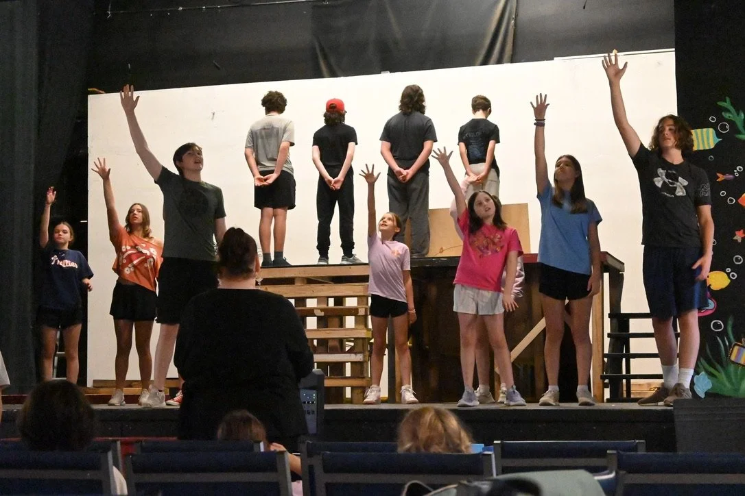 Children performing on a stage with some standing on the floor and some on a raised platform, audience watching.