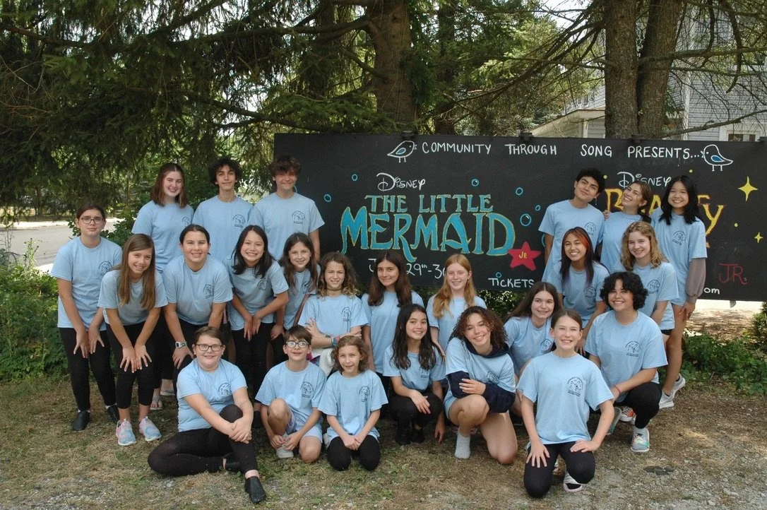 A group of children and teenagers posing outdoors in front of a sign for "The Little Mermaid" musical. They are all wearing matching light blue T-shirts.