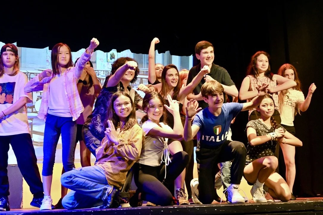Group of young people on stage, smiling and posing with some raising their fists or hands, in front of a painted backdrop, during a performance or rehearsal.