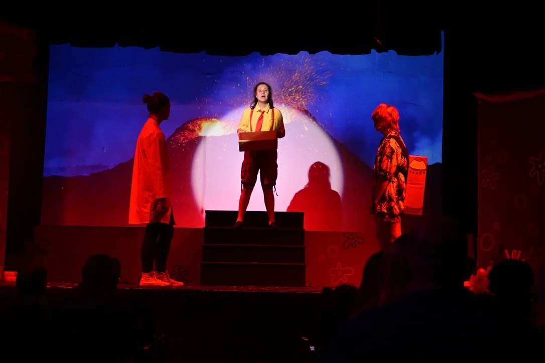 Three children performing on stage with a volcano eruption background. One girl in yellow and red standing on the steps, holding a box. Two other kids, one in red and the other with red hair and a patterned shirt, standing on each side.