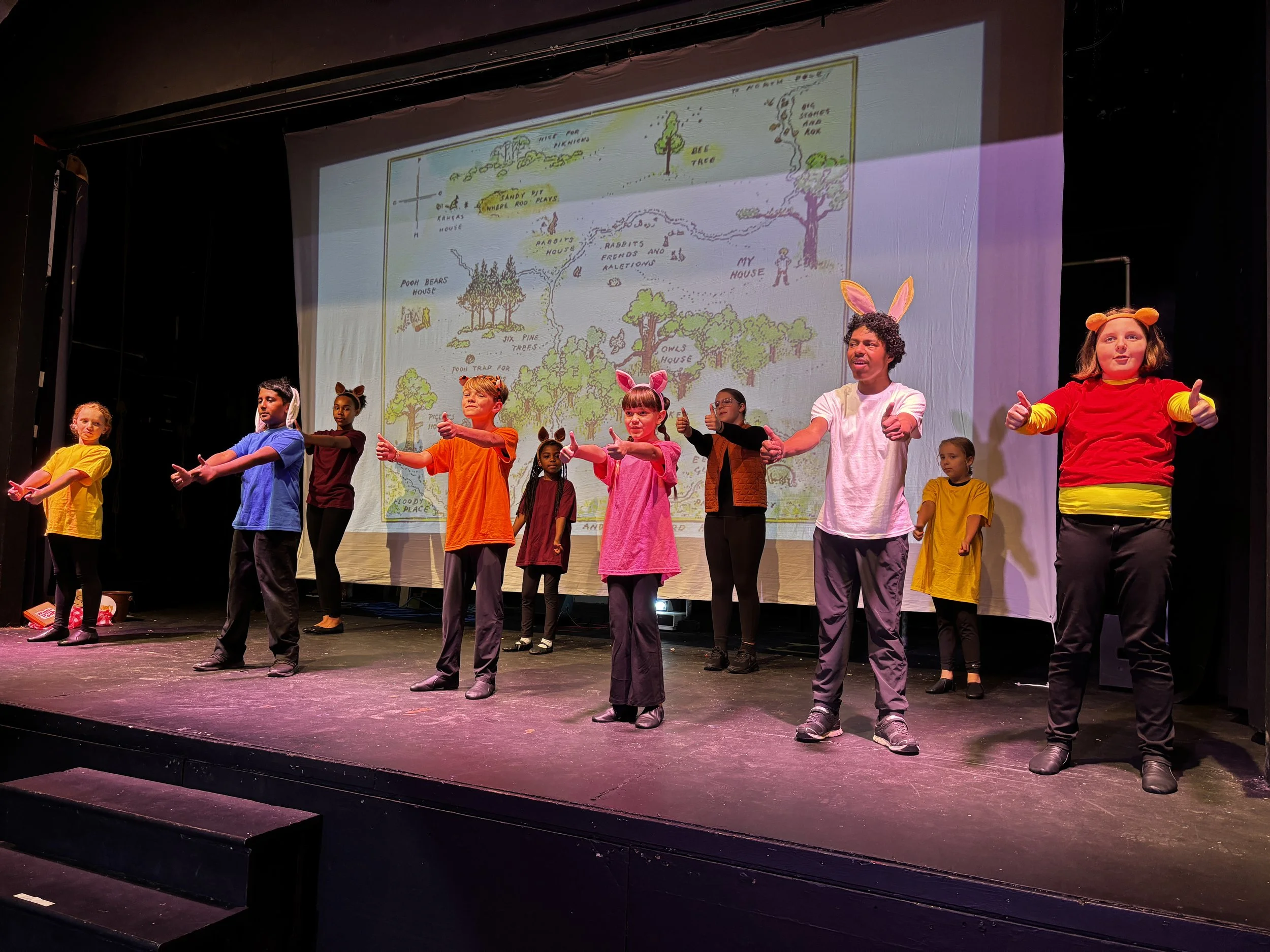 Children in colorful costumes performing on stage with a large illustrated backdrop of a forest scene.