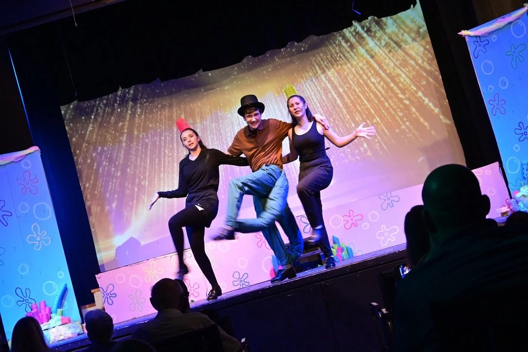 Three performers on stage in front of a colorful, animated background, engaging in a dance or theatrical performance. The audience is seated and watching the show.