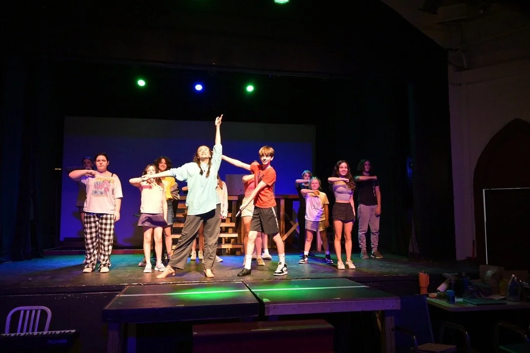Children performing on stage during a play or rehearsal, with some in colorful casual clothing and focused expressions.