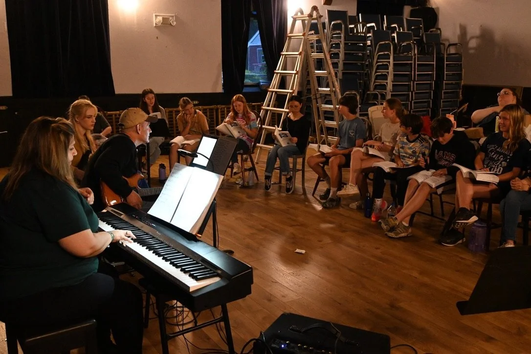 A music rehearsal or class with a woman playing piano, a man with a guitar, and children sitting in chairs with books, in a room with a stage, stacked chairs, and ladders.