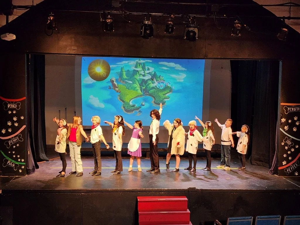 Children performing on stage in a play with a backdrop of a fantasy island surrounded by water and a sunny sky.