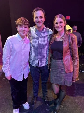 Three people standing together in a dark indoor setting, smiling for the camera. The person on the left is a young male with curly hair, wearing a white shirt and dark pants. The person in the middle is an adult male with short dark hair, wearing a s