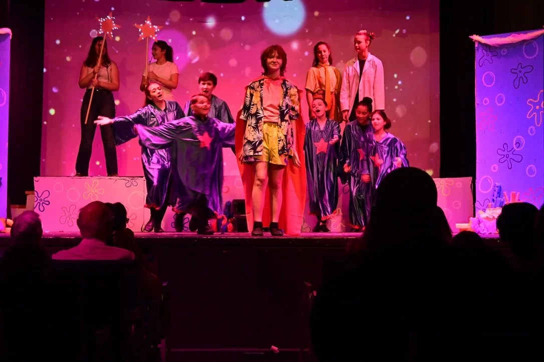 Children performing on stage in colorful costumes, with a backdrop featuring pink and purple hues and whimsical patterns, during a school play or talent show.