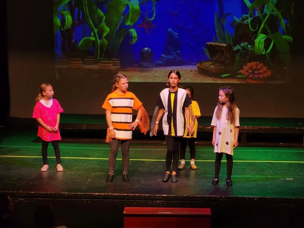 Five children are on stage performing a play with a colorful underwater scene featuring plants, a starfish, a treasure chest, and marine life projected in the background.