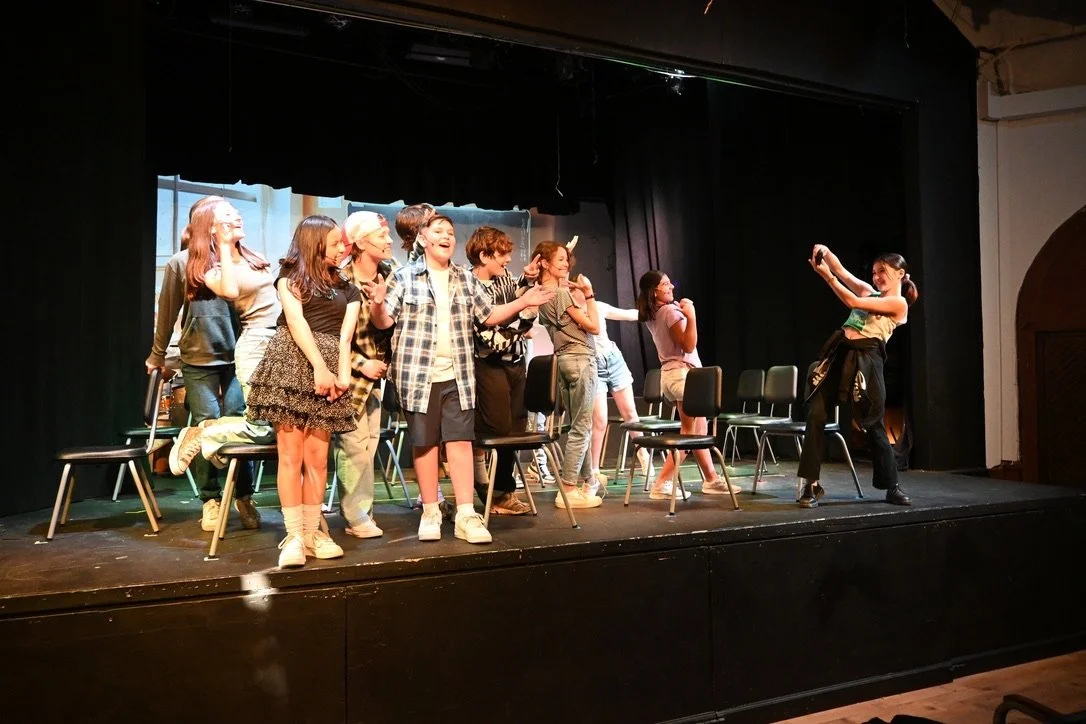 Children performing a play or skit on stage with chairs, some smiling, and one girl taking a photo or video of the group.