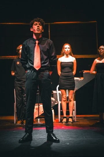 Young man in black dress shirt and pink tie performing on stage with three women standing behind him, all dressed in black, in a theatrical setting.