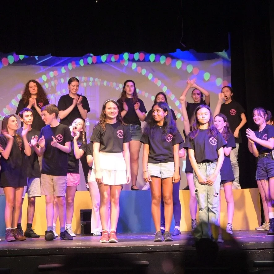 Group of children and teenagers on stage during a performance, some are standing on a colorful platform, wearing black t-shirts with a logo, with a colorful light display in the background.