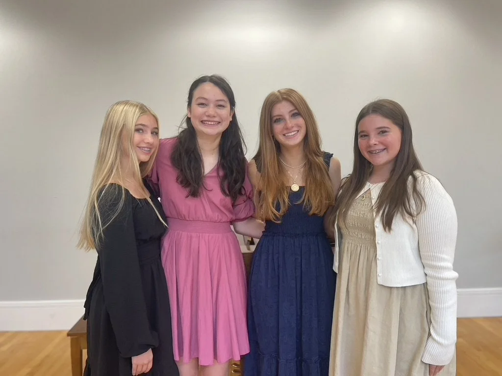 Four girls standing together indoors, smiling, with plain wall background and wooden floor, dressed in casual and semi-formal clothes.