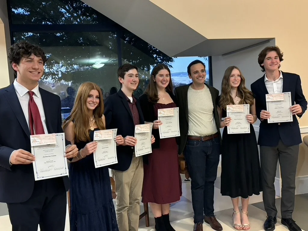 Seven young adults and an older man standing inside, holding certificates, smiling for a group photo at a celebration or awards event.