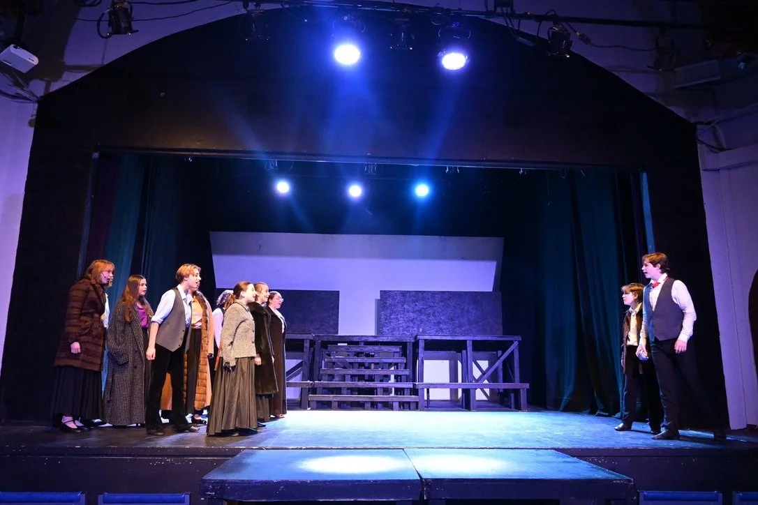 Theater stage with actors in vintage costumes facing each other, stage set with stairs and minimal scenery, lit by blue stage lights.