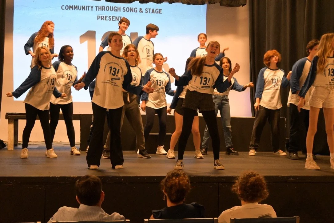 Group of young performers on stage, dancing during a community event, with a large screen behind them displaying event details.