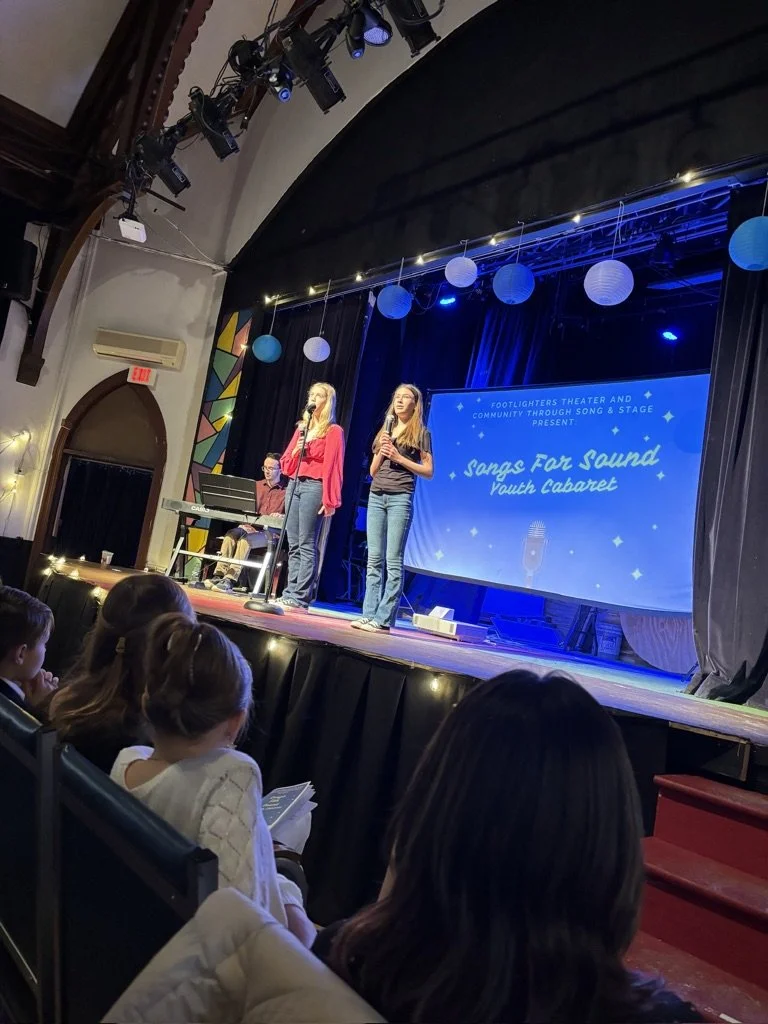 Two young women are singing on stage with a keyboard player in a theater. The stage has a large screen with the words 'Songs For Sound Youth Cabaret' and musical notes, and it is decorated with hanging paper lanterns. Audience members are seated, wat