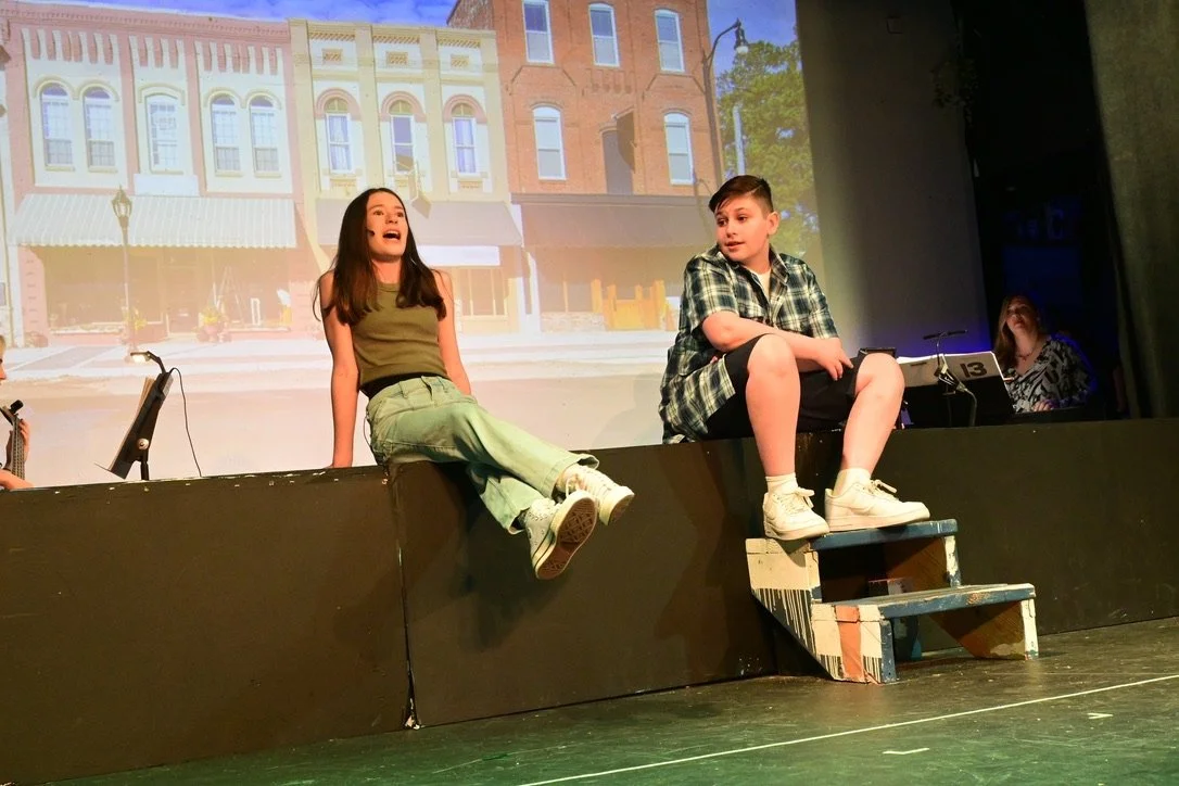 Two children, a girl and a boy, are sitting on a stage ledge, performing in a play with a storefront backdrop projected on the wall behind them. The girl is speaking, while the boy is listening, with a small set of stairs beside him.