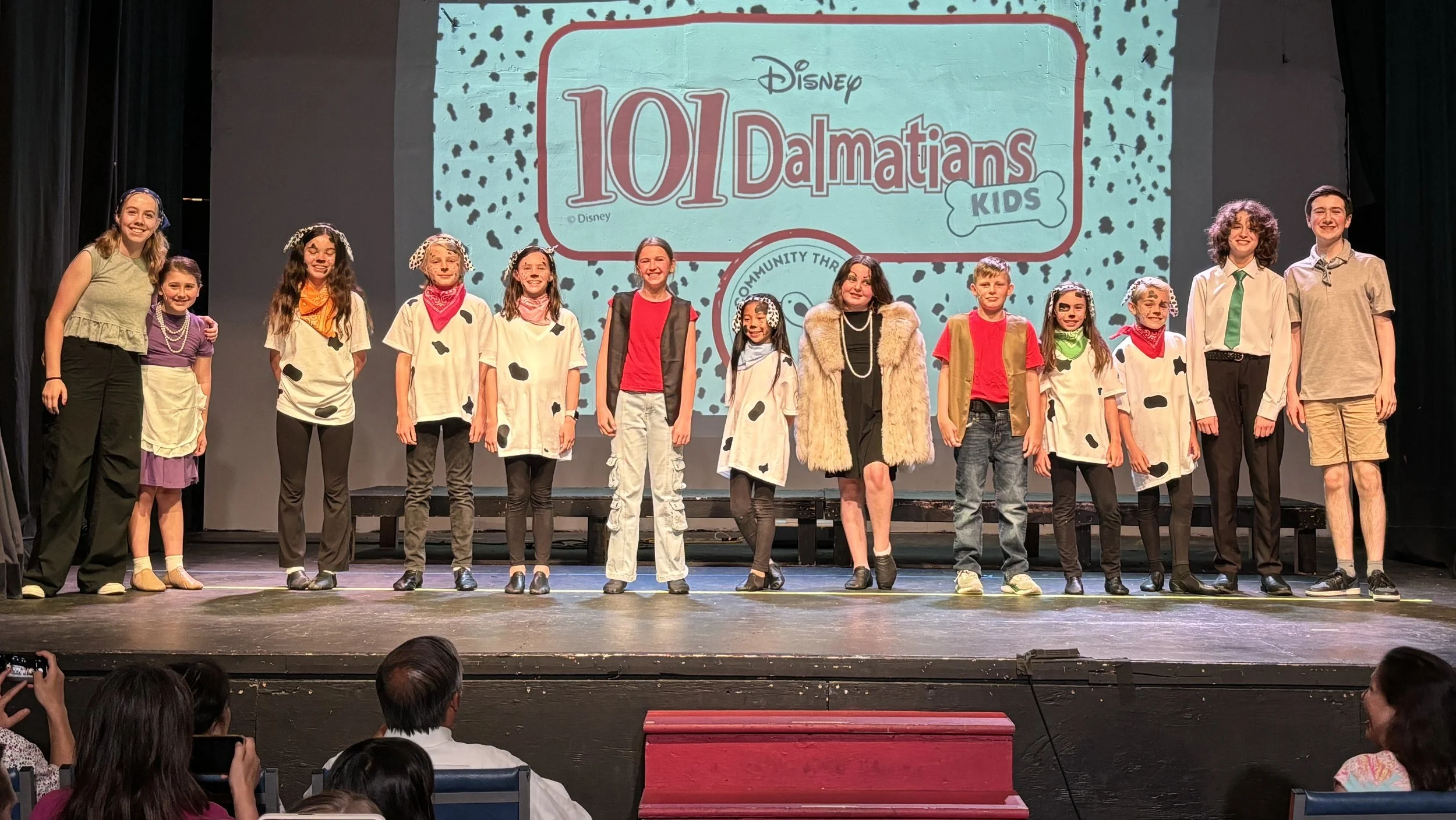 Children on stage dressed in costumes from the movie 101 Dalmatians during a performance, with an audience watching in front of the stage.