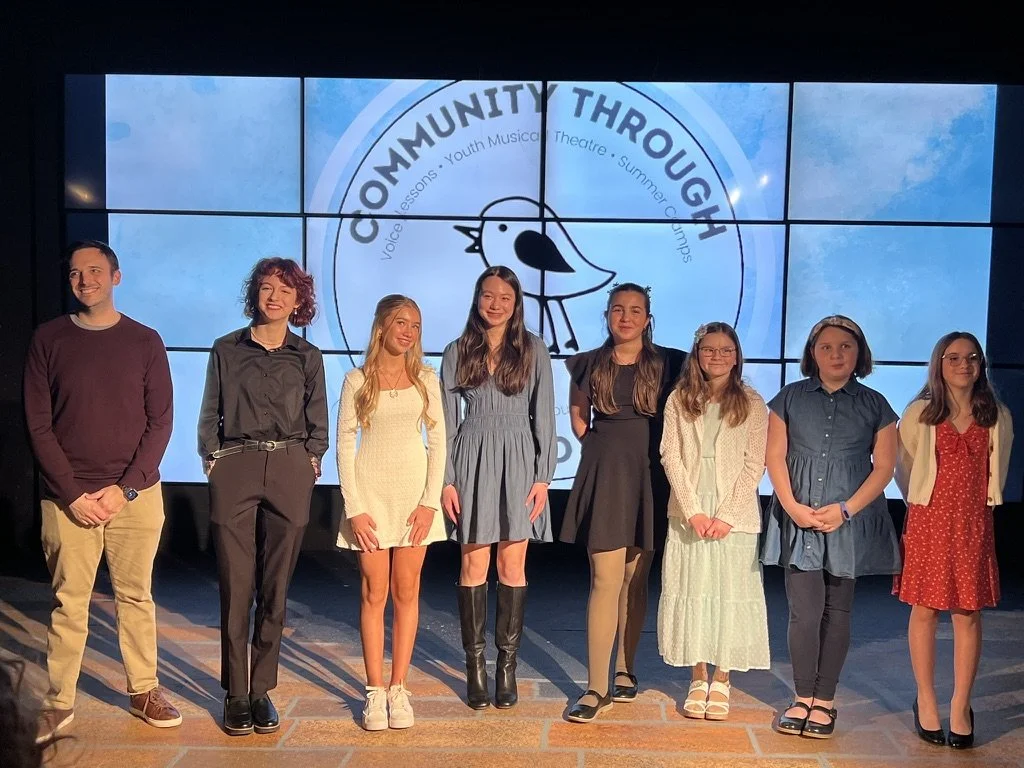 Group of eight young people standing on stage in front of a large screen displaying a logo for 'Community Through' with a cartoon bird and text about youth music, theater, and summer camp.