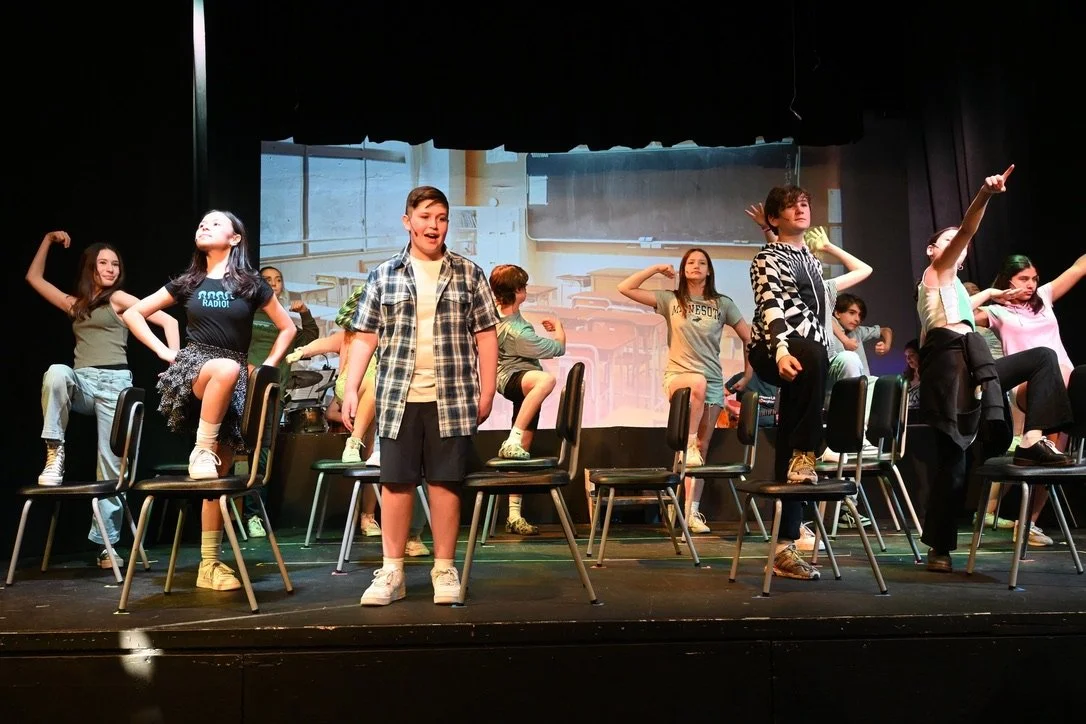 Children performing a school play on stage, with some standing on chairs and others singing or dancing in front of a classroom backdrop.