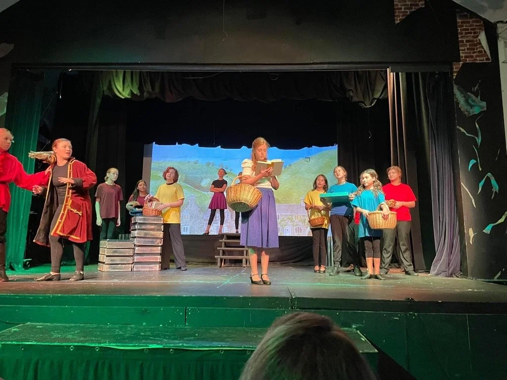 A group of children and teenagers performing a play or musical on stage, with some holding baskets and others reading from scripts, in front of a scenic backdrop.