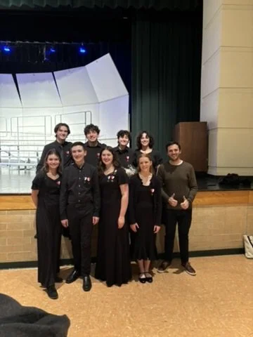 Group of nine young adults posing on stage, dressed in formal black attire, smiling and standing in front of a music stand and stage setup.