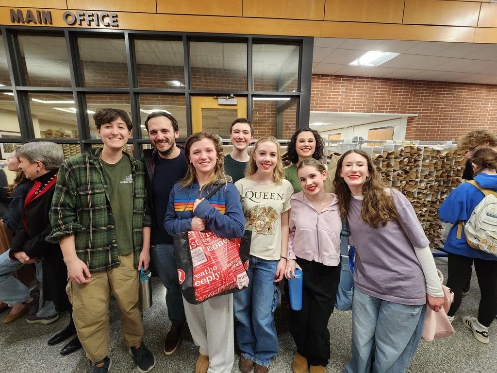 Group of eight smiling students and one adult standing in front of a school main office entrance, with other students and adults in the background.