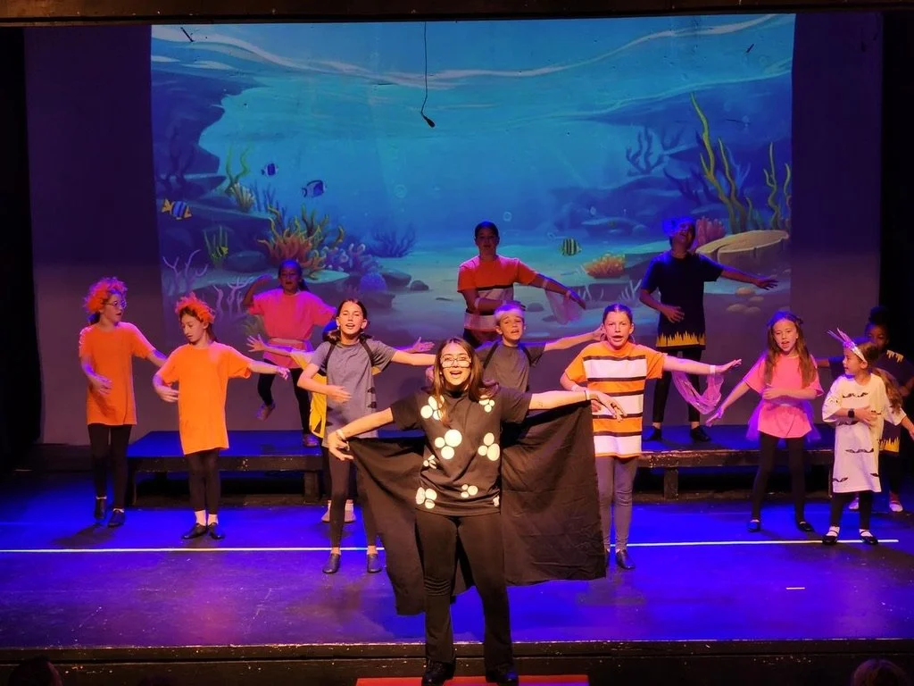 Group of children performing a play with an ocean scene projected on the background, some children dressed in colorful costumes and some with accessories like trophies or headbands.