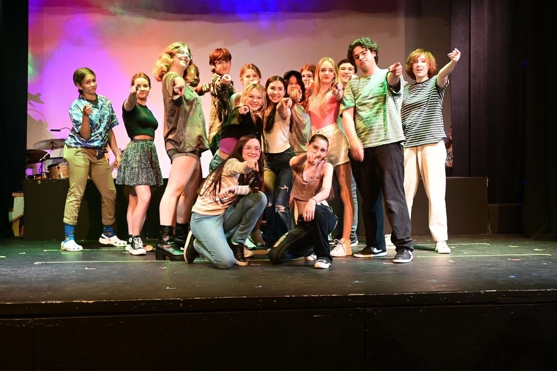 A group of young performers on stage, posing with enthusiasm after a performance, with colorful lighting and musical instruments in the background.