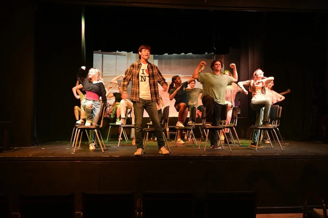 A group of young people performing on stage with chairs, some standing and some sitting, under stage lights, with a painted background behind them.