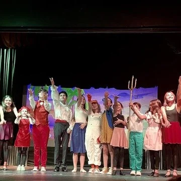 A group of children and teenagers dressed in festive costumes and casual clothes, standing on a stage with their arms raised in a celebratory gesture, during a performance or celebration.