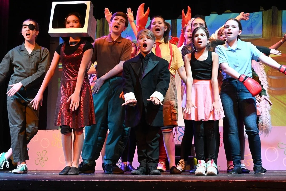 Children performing in a school play, some with costumes like crab claws and a TV head, on stage with colorful backdrop.