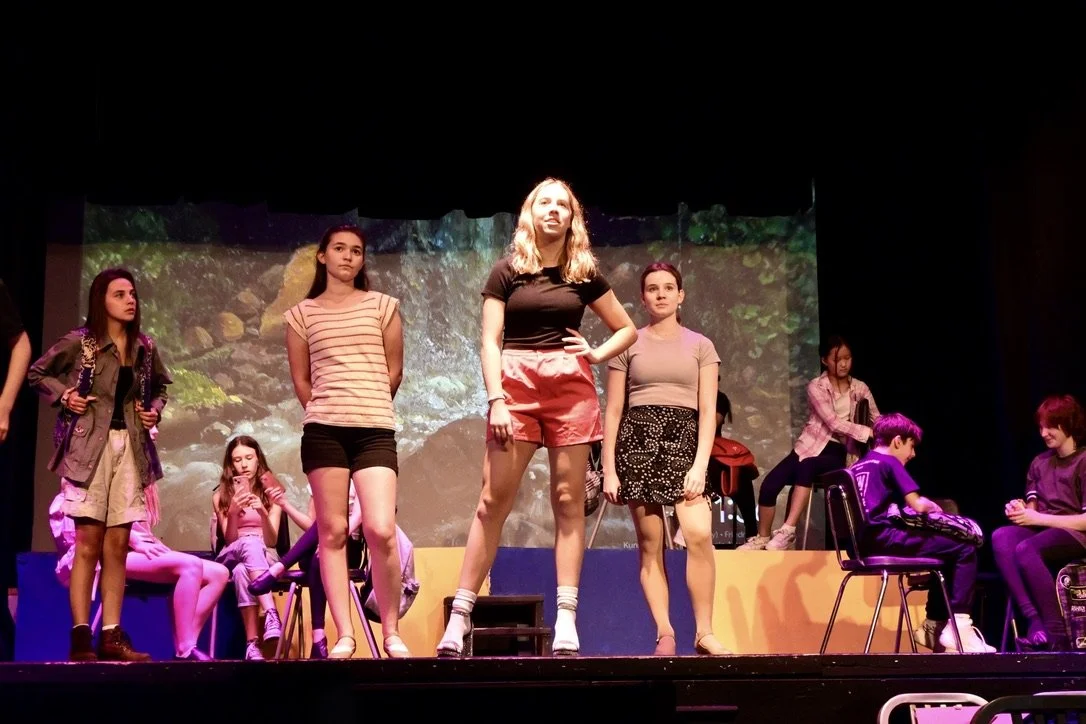 Group of young women on stage during a theater performance, with a scenic background of a forest or nature scene.