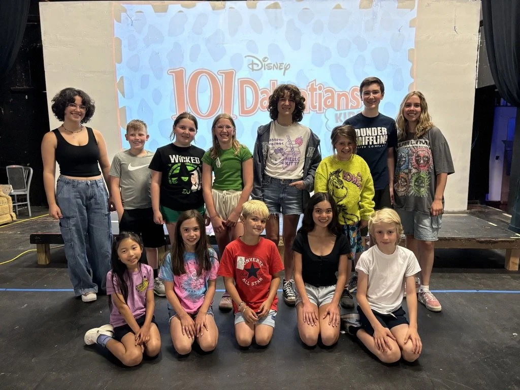 A group of fifteen children and teenagers posing on a stage in front of a screen that says "101 Dalmatians" with a Disney logo, some smiling and wearing colorful casual clothing.