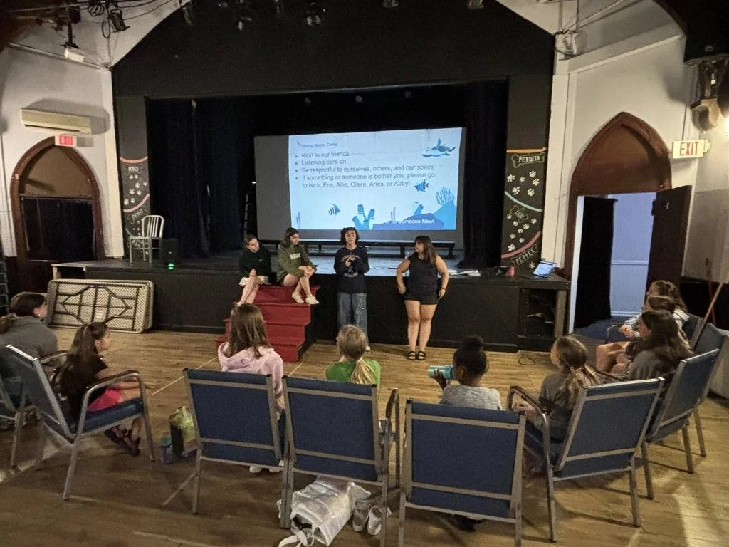 Children sitting in chairs watching a presentation on a stage with three women speaking. The slide on the screen has text about kindness, listening, and respect.