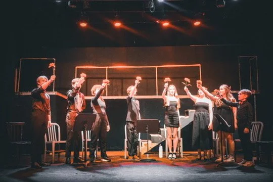 Theatre cast taking a bow on stage, holding hats, with chairs and props in the background.