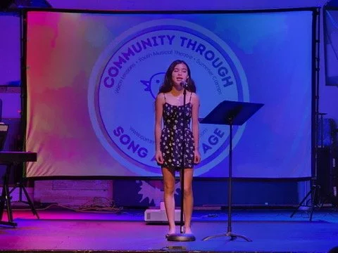 Young girl singing on stage with a microphone, in front of a screen displaying the logo for 'Community Through Song Age'.