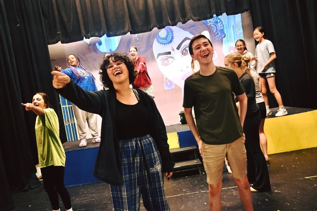 Group of children and teenagers laughing and acting on stage during a theater rehearsal or performance, with colorful set and backdrop.