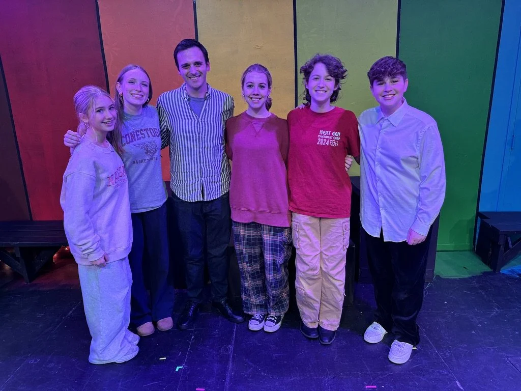 Group of six people, including kids and an adult, standing on stage in front of a colorful wooden block wall, smiling for a group photo.