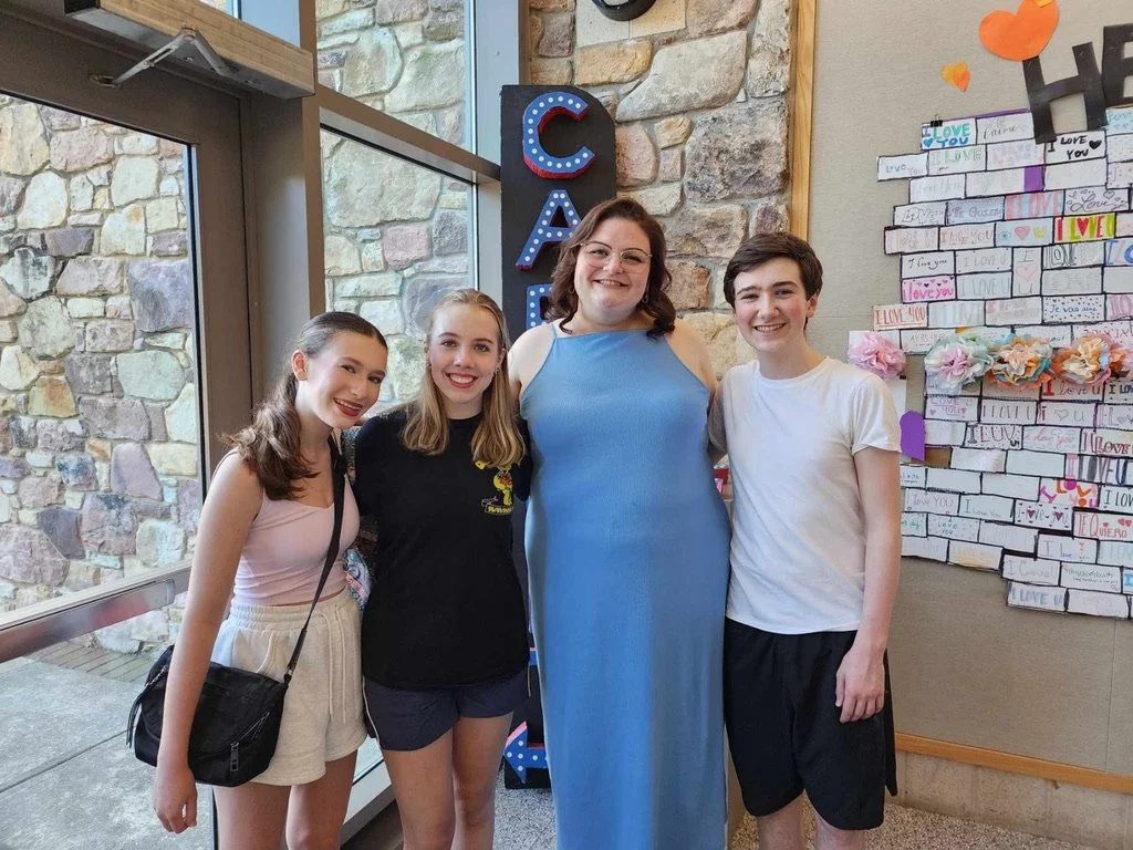 Four young people, three women and one man, smiling and standing together indoors near a decorated wall with notes and hearts, next to a large stone wall and glass door.