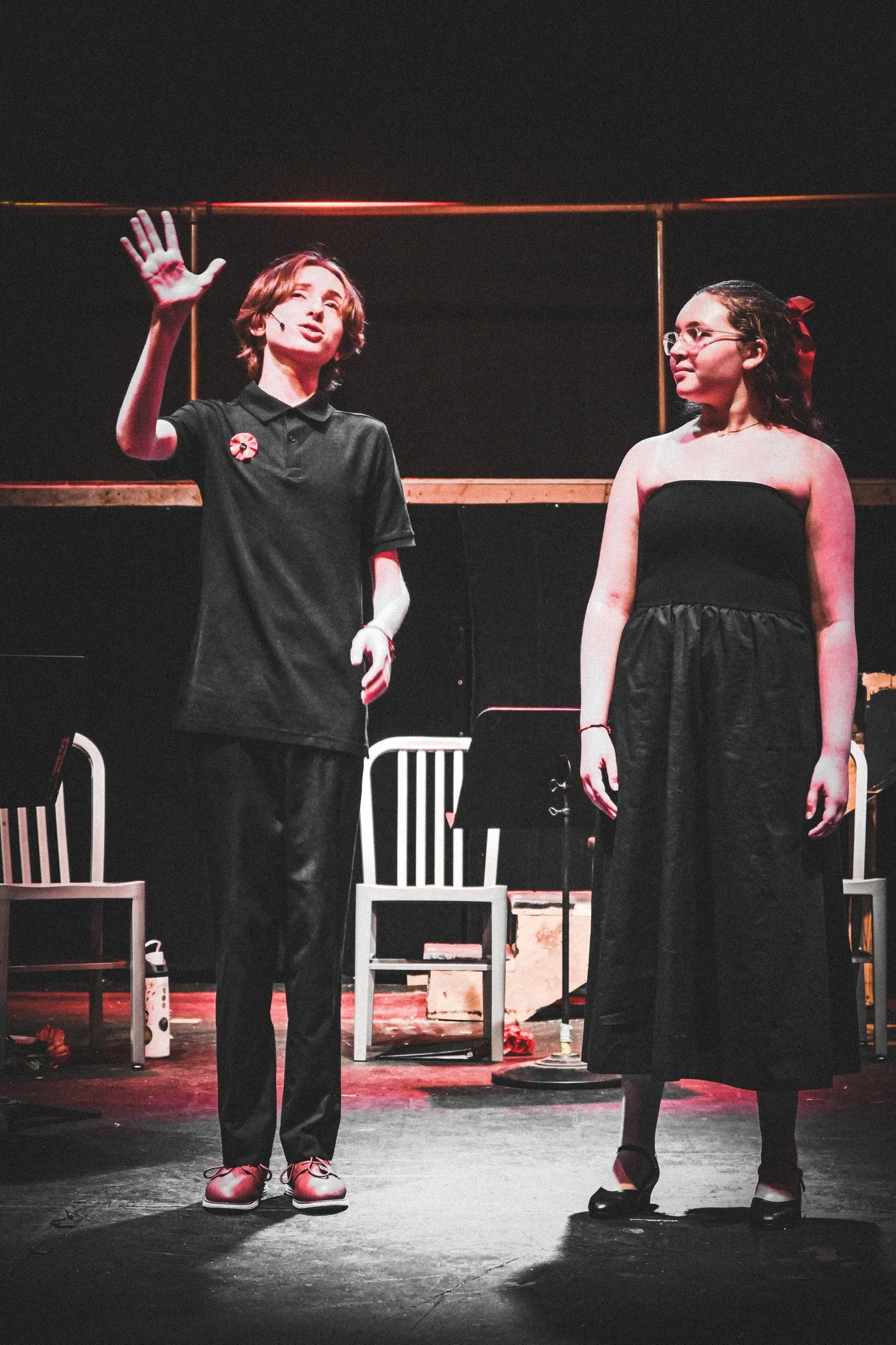 Two women standing on stage during a performance. One woman, on the left, has short brown hair and is wearing a black polo shirt, black pants, and brown shoes, and is raising her right hand. The other woman, on the right, has long, curly hair tied ba