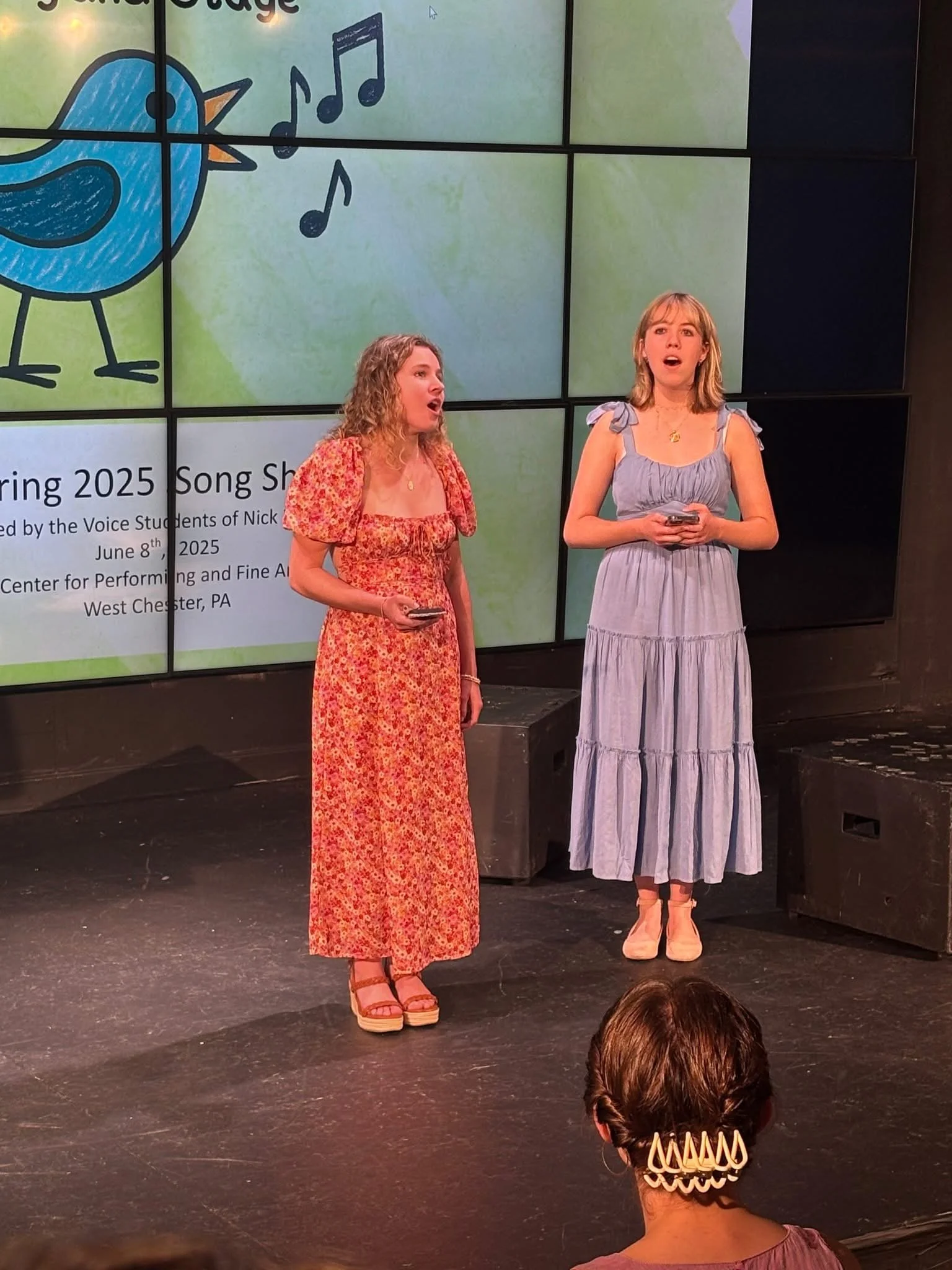 Two women standing on stage singing, with a large monitor behind them displaying singing notes, a cartoon bird, and event information for a spring 2025 song showcase.