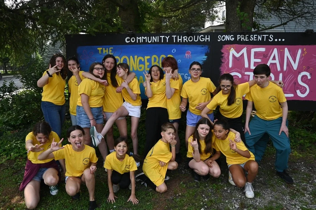 Group of children and teenagers in yellow T-shirts posing in front of a colorful backdrop with the text 'The SpongeBob' and 'Mean Girls' at an outdoor event.